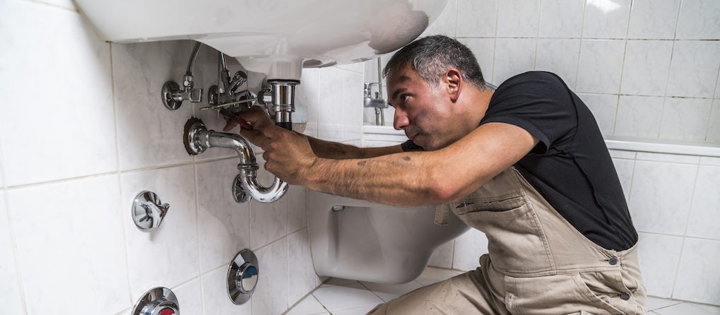 plumber in black t-shirt and white working suit repairs faucet pipe with a metallic pliers in bathroom.