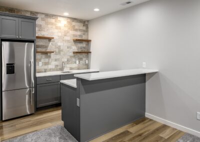 Modern kitchen with a stainless steel fridge, gray cabinets, stone backsplash, and a white countertop island. The blend of wood and carpet flooring complements the sleek design.