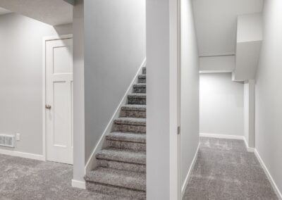 A modern, minimalistic hallway with gray carpeted stairs on the left leading to a basement. A narrow passageway on the right guides you to a brightly lit area. Walls and ceiling are painted white.