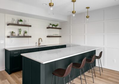 Recently remodeled, this modern kitchen boasts a large island with white countertops and dark green cabinets. Four brown bar stools provide seating. Open shelves adorned with plants and glassware decorate the wall, while pendant lights elegantly hang from the ceiling.