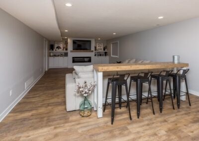 Remodeled basement room with a wooden floor, featuring a bar table with stools, a beige sectional sofa, and a wall-mounted TV with cabinets.