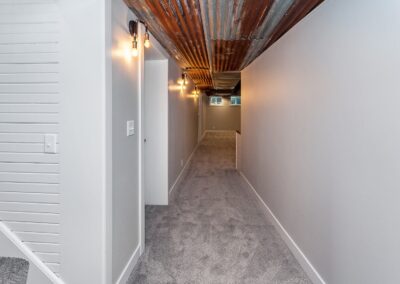 A narrow basement hallway with a gray carpet and corrugated metal ceiling, featuring exposed bulb lighting.