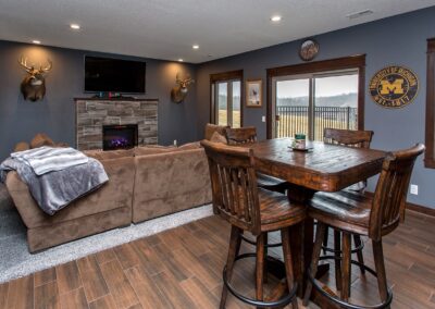 The living room features brown couches, a stone fireplace, mounted deer heads, a wall-mounted TV, and a high wooden table with chairs. Wall decor includes a University of Michigan emblem—ideal for any handyman to gather after tackling that bathroom remodel project.