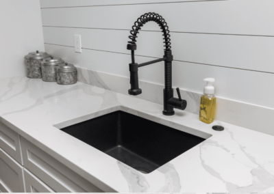 A modern kitchen remodel features a sleek sink with a flexible black faucet set in a white marble countertop. Nearby, three metal canisters and a soap dispenser add charm, while the white shiplap wall provides a crisp backdrop.