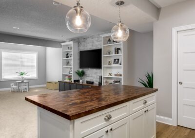 Modern living room with a wooden island counter, gray and white decor, shelving units, and a wall-mounted TV, recently remodeled for style and function. A separate play area is visible in the background.