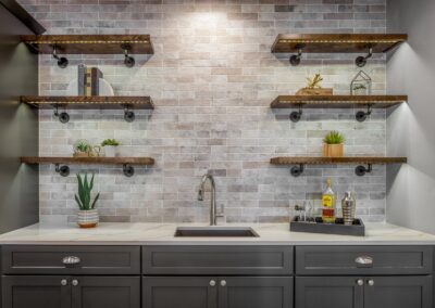 A modern kitchen with a tiled backsplash, wooden shelves holding various objects, and a sleek silver faucet gracing the sink. The counter is adorned with plants and a tray of condiments, creating an inviting basement vibe.