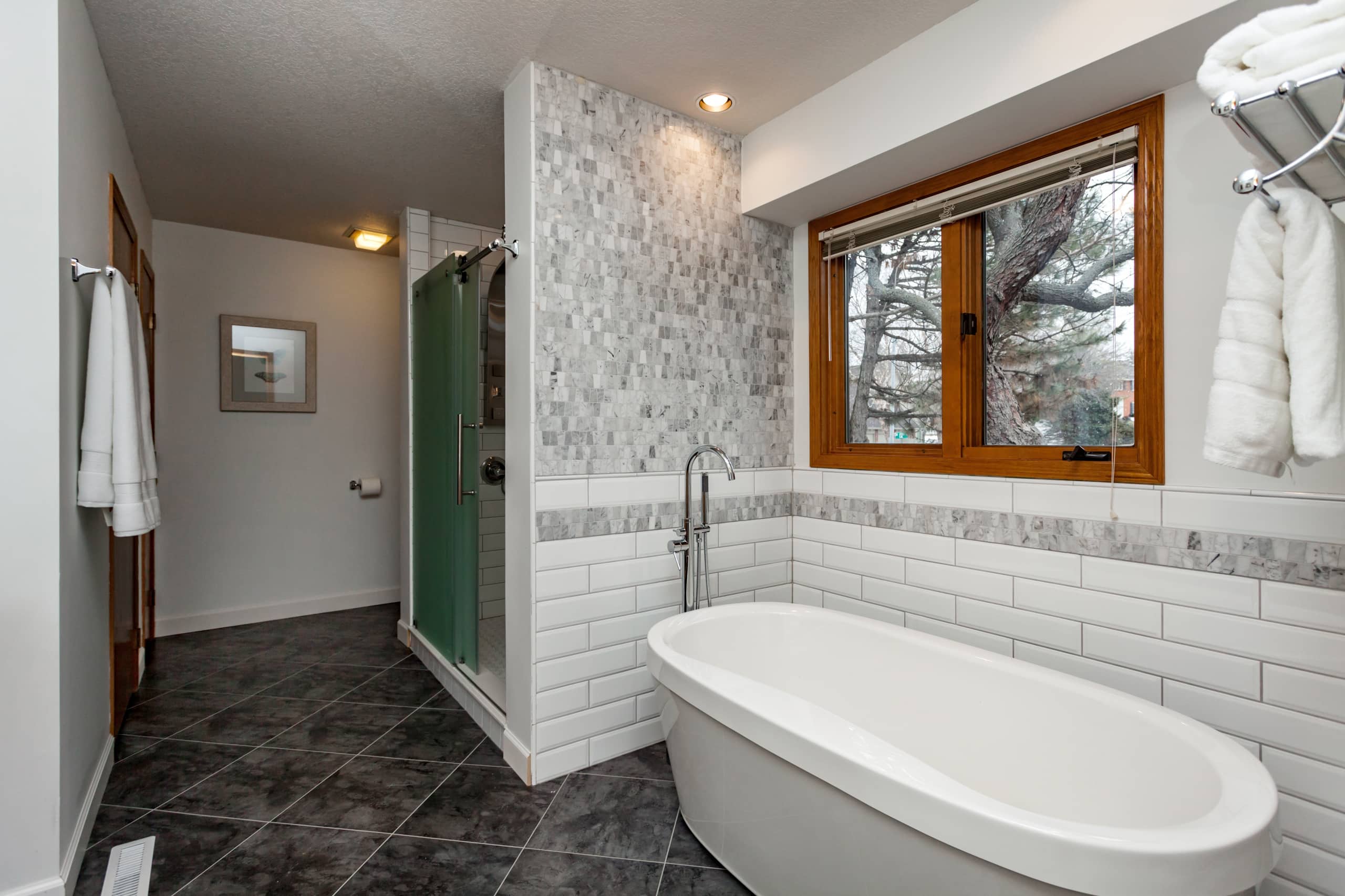img-24 A modern bathroom features a freestanding bathtub, glass-enclosed shower, and gray tile flooring. White subway tiles line the walls, while a window bathes the space in natural light—creating an inviting sanctuary that even a skilled handyman would appreciate for its meticulous craftsmanship.